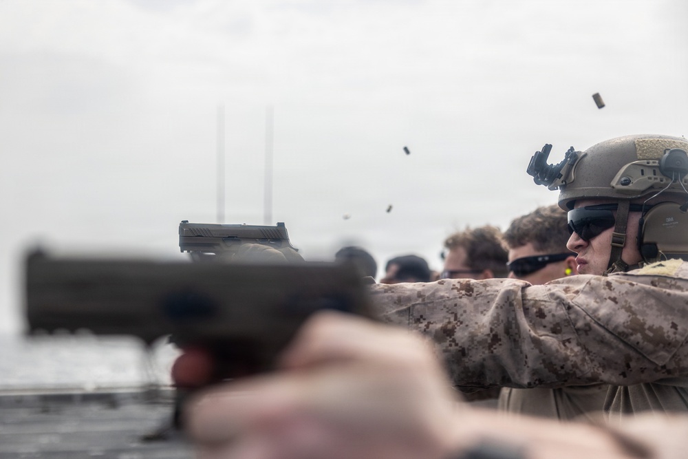 Military personnel, appearing to be U.S. Marines based on MARPAT camouflage uniforms, are conducting a live-fire pistol qualification or training exercise on what appears to be a ship deck. Multiple spent brass casings are visibly ejecting from the firearms mid-shot, captured in motion. Personnel are equipped with tactical helmets, hearing protection, and eye protection.