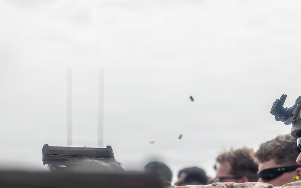 Military personnel, appearing to be U.S. Marines based on MARPAT camouflage uniforms, are conducting a live-fire pistol qualification or training exercise on what appears to be a ship deck. Multiple spent brass casings are visibly ejecting from the firearms mid-shot, captured in motion. Personnel are equipped with tactical helmets, hearing protection, and eye protection.