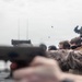 Military personnel, appearing to be U.S. Marines based on MARPAT camouflage uniforms, are conducting a live-fire pistol qualification or training exercise on what appears to be a ship deck. Multiple spent brass casings are visibly ejecting from the firearms mid-shot, captured in motion. Personnel are equipped with tactical helmets, hearing protection, and eye protection.