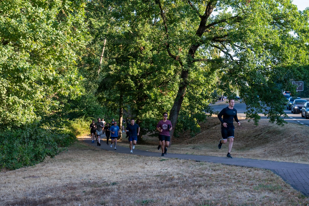 A group of runners jogs along a paved path through a park-like setting with large green trees. The scene appears to be a group fitness run or informal race, with approximately 10-12 participants spread across the path. The setting suggests a warm, dry summer day based on the parched grass and bright sunlight.