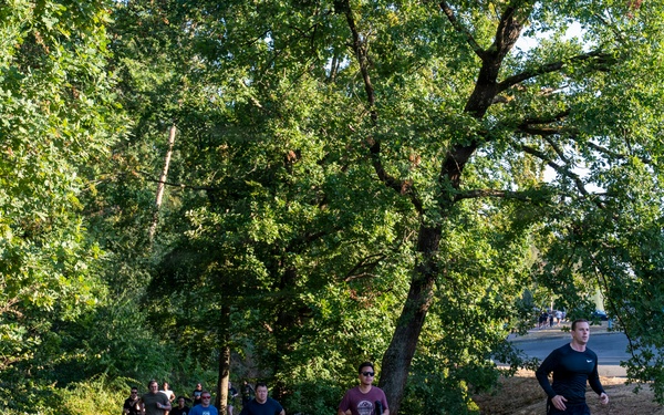 A group of runners jogs along a paved path through a park-like setting with large green trees. The scene appears to be a group fitness run or informal race, with approximately 10-12 participants spread across the path. The setting suggests a warm, dry summer day based on the parched grass and bright sunlight.