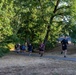 A group of runners jogs along a paved path through a park-like setting with large green trees. The scene appears to be a group fitness run or informal race, with approximately 10-12 participants spread across the path. The setting suggests a warm, dry summer day based on the parched grass and bright sunlight.