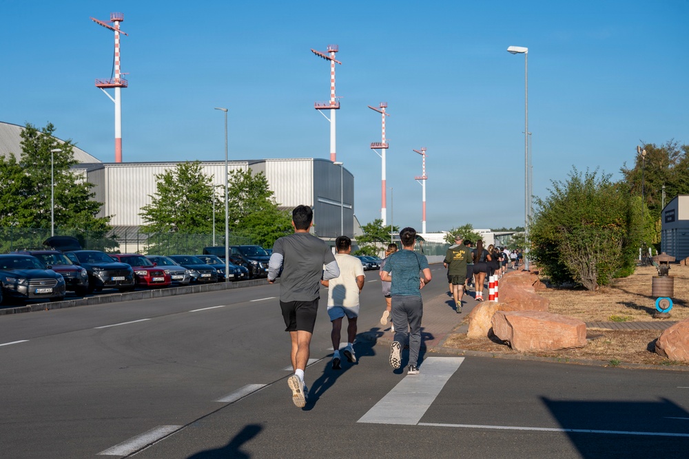 A group of people are running along a road in what appears to be an industrial or airport-adjacent area, with red-and-white striped navigation or obstruction marker poles visible in the background. The scene is captured from behind the runners, showing a line of participants extending into the distance. The setting includes a parking lot on the left, large industrial buildings, and landscaping with rocks and shrubs on the right.