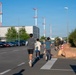 A group of people are running along a road in what appears to be an industrial or airport-adjacent area, with red-and-white striped navigation or obstruction marker poles visible in the background. The scene is captured from behind the runners, showing a line of participants extending into the distance. The setting includes a parking lot on the left, large industrial buildings, and landscaping with rocks and shrubs on the right.
