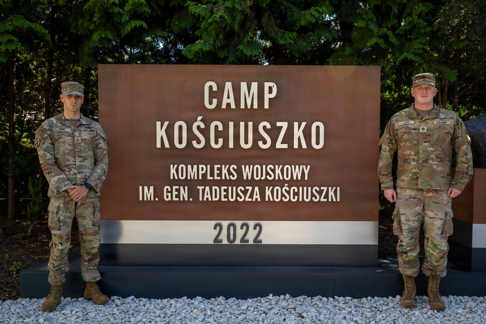 Two U.S. Army soldiers in MultiCam (OCP) uniforms stand on either side of a large entrance sign for Camp Kościuszko, a military installation named after General Tadeusz Kościuszko, established in 2022. The sign is bilingual, displaying both English and Polish text. The setting appears to be in Poland, consistent with the Polish language subtitle 'Kompleks Wojskowy Im. Gen. Tadeusza Kościuszki'.
