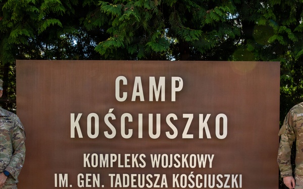Two U.S. Army soldiers in MultiCam (OCP) uniforms stand on either side of a large entrance sign for Camp Kościuszko, a military installation named after General Tadeusz Kościuszko, established in 2022. The sign is bilingual, displaying both English and Polish text. The setting appears to be in Poland, consistent with the Polish language subtitle 'Kompleks Wojskowy Im. Gen. Tadeusza Kościuszki'.