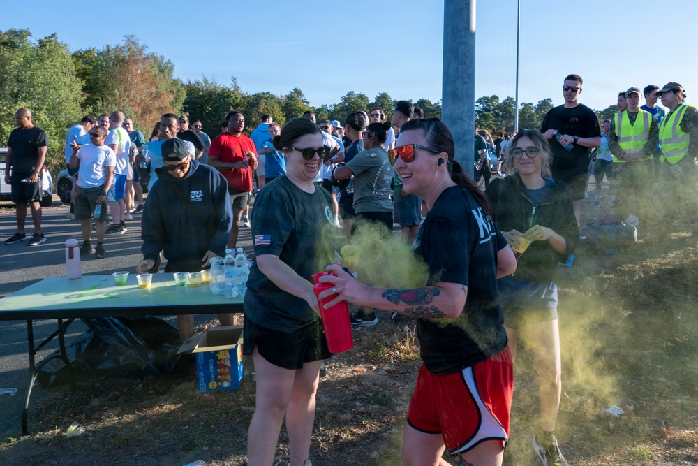 An outdoor color run or fun run event with a large crowd of participants gathered near a refreshment table. Two women in the foreground are the focal point, with one spraying yellow/green colored powder from a red container. The event appears to be taking place on a sunny day in a parking lot or road area bordered by trees.