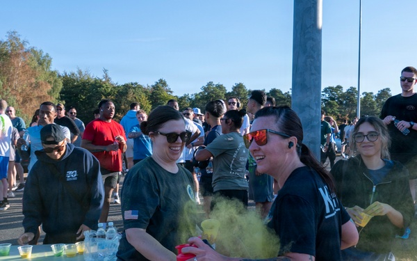An outdoor color run or fun run event with a large crowd of participants gathered near a refreshment table. Two women in the foreground are the focal point, with one spraying yellow/green colored powder from a red container. The event appears to be taking place on a sunny day in a parking lot or road area bordered by trees.