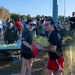 An outdoor color run or fun run event with a large crowd of participants gathered near a refreshment table. Two women in the foreground are the focal point, with one spraying yellow/green colored powder from a red container. The event appears to be taking place on a sunny day in a parking lot or road area bordered by trees.