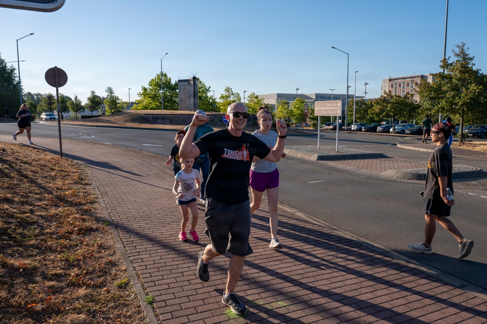 A group of people are running or jogging along a brick sidewalk in what appears to be an urban or suburban area, likely during a fun run or community race event. The central figure is a bald man wearing a black 'Tough Mudder' t-shirt with his fist raised in a celebratory gesture. A young child in pink shoes and several adults are visible participating in the activity.