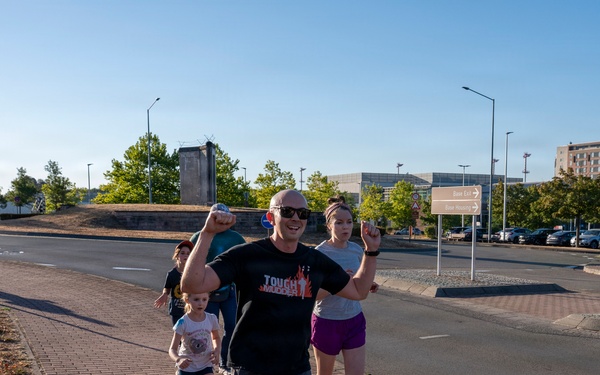 A group of people are running or jogging along a brick sidewalk in what appears to be an urban or suburban area, likely during a fun run or community race event. The central figure is a bald man wearing a black 'Tough Mudder' t-shirt with his fist raised in a celebratory gesture. A young child in pink shoes and several adults are visible participating in the activity.