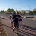 A group of people are running or jogging along a brick sidewalk in what appears to be an urban or suburban area, likely during a fun run or community race event. The central figure is a bald man wearing a black 'Tough Mudder' t-shirt with his fist raised in a celebratory gesture. A young child in pink shoes and several adults are visible participating in the activity.
