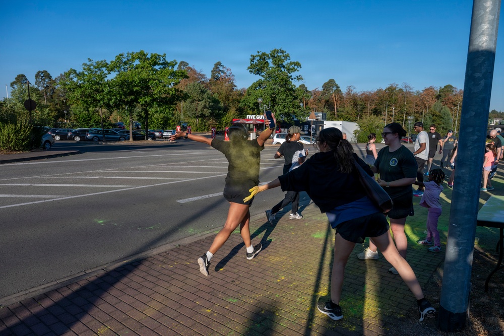 An outdoor color run or powder paint event is taking place in a paved area near a parking lot. Participants are throwing or applying colored powder (primarily yellow/green) at each other in a festive, energetic atmosphere. A Five Guys restaurant sign is visible in the background, suggesting this may be on or near a commercial or military base.