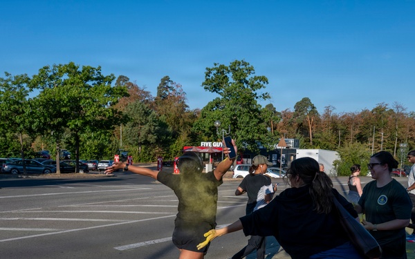 An outdoor color run or powder paint event is taking place in a paved area near a parking lot. Participants are throwing or applying colored powder (primarily yellow/green) at each other in a festive, energetic atmosphere. A Five Guys restaurant sign is visible in the background, suggesting this may be on or near a commercial or military base.