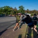 An outdoor color run or powder paint event is taking place in a paved area near a parking lot. Participants are throwing or applying colored powder (primarily yellow/green) at each other in a festive, energetic atmosphere. A Five Guys restaurant sign is visible in the background, suggesting this may be on or near a commercial or military base.