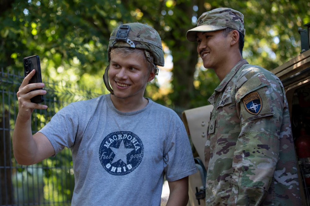 A civilian wearing a 'Macedonia Ohrid' t-shirt and a military helmet is taking a selfie with a U.S. Army soldier in full multicam uniform. The soldier displays a NATO patch and a U.S. Army tape on his uniform, suggesting a NATO-affiliated deployment. Both individuals are smiling, indicating a friendly, informal interaction.