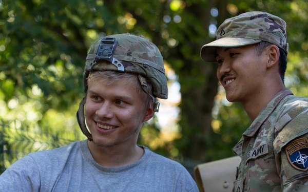 A civilian wearing a 'Macedonia Ohrid' t-shirt and a military helmet is taking a selfie with a U.S. Army soldier in full multicam uniform. The soldier displays a NATO patch and a U.S. Army tape on his uniform, suggesting a NATO-affiliated deployment. Both individuals are smiling, indicating a friendly, informal interaction.