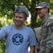 A civilian wearing a 'Macedonia Ohrid' t-shirt and a military helmet is taking a selfie with a U.S. Army soldier in full multicam uniform. The soldier displays a NATO patch and a U.S. Army tape on his uniform, suggesting a NATO-affiliated deployment. Both individuals are smiling, indicating a friendly, informal interaction.