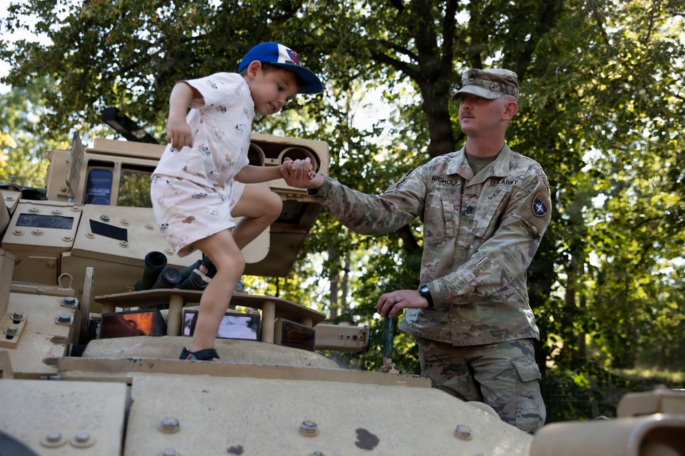 A U.S. Army soldier in OCP (Operational Camouflage Pattern) uniform assists a young child climbing on top of a tan military vehicle, likely a Humvee or similar armored vehicle. The soldier, whose name tape reads 'NICHOLS' and wears a U.S. Army patch, holds the child's hand to steady them. The setting appears to be an outdoor public event, possibly a military open house or community outreach event, with trees visible in the background.