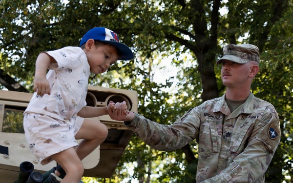 A U.S. Army soldier in OCP (Operational Camouflage Pattern) uniform assists a young child climbing on top of a tan military vehicle, likely a Humvee or similar armored vehicle. The soldier, whose name tape reads 'NICHOLS' and wears a U.S. Army patch, holds the child's hand to steady them. The setting appears to be an outdoor public event, possibly a military open house or community outreach event, with trees visible in the background.