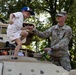 A U.S. Army soldier in OCP (Operational Camouflage Pattern) uniform assists a young child climbing on top of a tan military vehicle, likely a Humvee or similar armored vehicle. The soldier, whose name tape reads 'NICHOLS' and wears a U.S. Army patch, holds the child's hand to steady them. The setting appears to be an outdoor public event, possibly a military open house or community outreach event, with trees visible in the background.