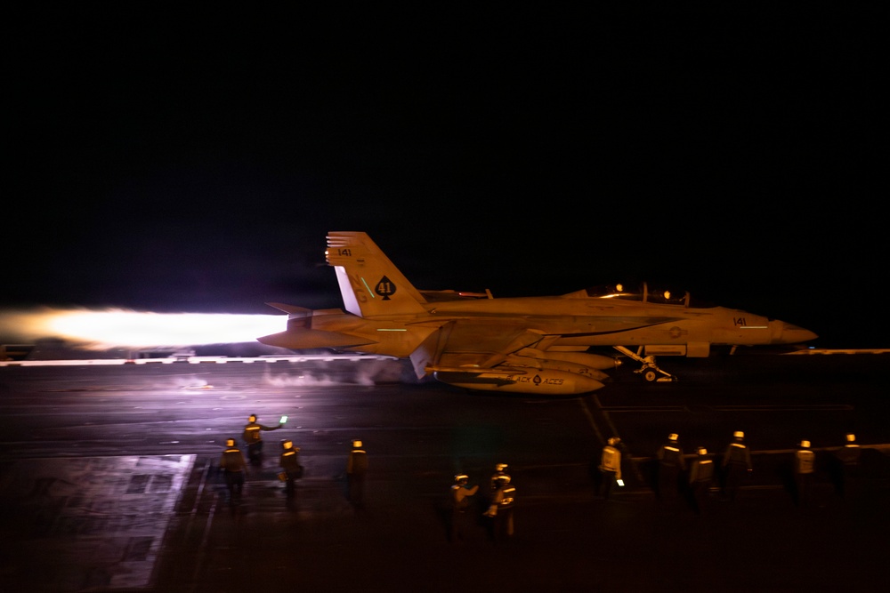 A military jet aircraft, marked with the number '141', is photographed at night on what appears to be an aircraft carrier flight deck. The aircraft's engines are producing visible afterburner exhaust, illuminating the wet deck surface. Multiple flight deck crew members in reflective gear are visible surrounding the aircraft.