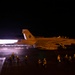 A military jet aircraft, marked with the number '141', is photographed at night on what appears to be an aircraft carrier flight deck. The aircraft's engines are producing visible afterburner exhaust, illuminating the wet deck surface. Multiple flight deck crew members in reflective gear are visible surrounding the aircraft.