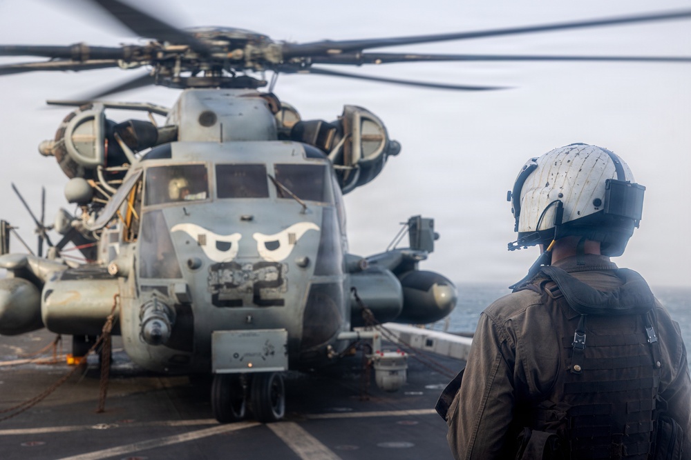 A military personnel wearing a white flight deck helmet and tactical gear stands on a ship's flight deck observing a large military helicopter, identifiable as a CH-53E Super Stallion, with distinctive eye-shaped nose art painted on its fuselage. The helicopter's rotors are spinning, suggesting it is either preparing for takeoff or has just landed. The setting appears to be aboard a naval vessel at sea, with overcast skies visible in the background.