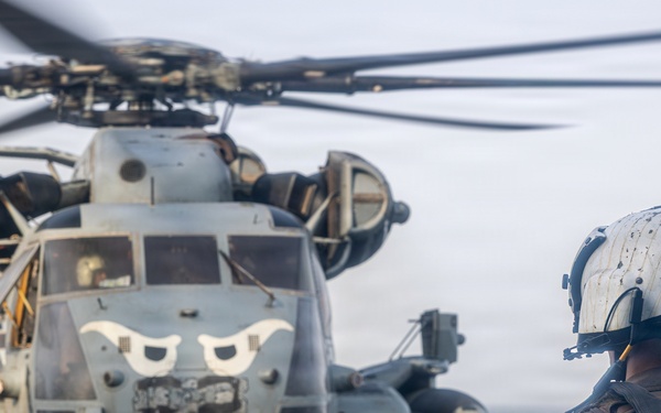 A military personnel wearing a white flight deck helmet and tactical gear stands on a ship's flight deck observing a large military helicopter, identifiable as a CH-53E Super Stallion, with distinctive eye-shaped nose art painted on its fuselage. The helicopter's rotors are spinning, suggesting it is either preparing for takeoff or has just landed. The setting appears to be aboard a naval vessel at sea, with overcast skies visible in the background.