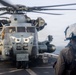 A military personnel wearing a white flight deck helmet and tactical gear stands on a ship's flight deck observing a large military helicopter, identifiable as a CH-53E Super Stallion, with distinctive eye-shaped nose art painted on its fuselage. The helicopter's rotors are spinning, suggesting it is either preparing for takeoff or has just landed. The setting appears to be aboard a naval vessel at sea, with overcast skies visible in the background.