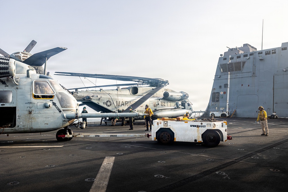 The image shows a naval vessel flight deck with U.S. Marine Corps helicopters being repositioned by deck crew. Multiple CH-53 Sea Stallion helicopters with folded rotor blades are visible, along with ground support equipment including a tow tractor. Deck crew members wearing yellow jerseys are actively working to maneuver the aircraft.