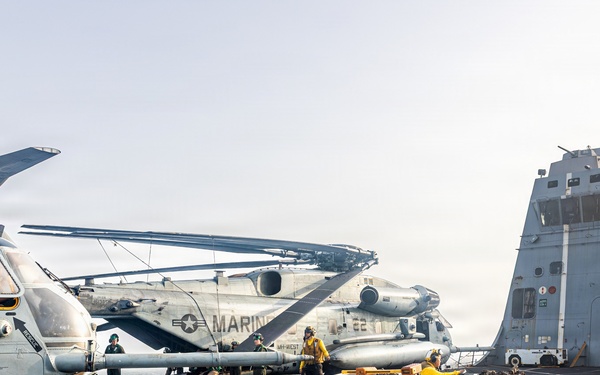 The image shows a naval vessel flight deck with U.S. Marine Corps helicopters being repositioned by deck crew. Multiple CH-53 Sea Stallion helicopters with folded rotor blades are visible, along with ground support equipment including a tow tractor. Deck crew members wearing yellow jerseys are actively working to maneuver the aircraft.