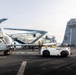 The image shows a naval vessel flight deck with U.S. Marine Corps helicopters being repositioned by deck crew. Multiple CH-53 Sea Stallion helicopters with folded rotor blades are visible, along with ground support equipment including a tow tractor. Deck crew members wearing yellow jerseys are actively working to maneuver the aircraft.