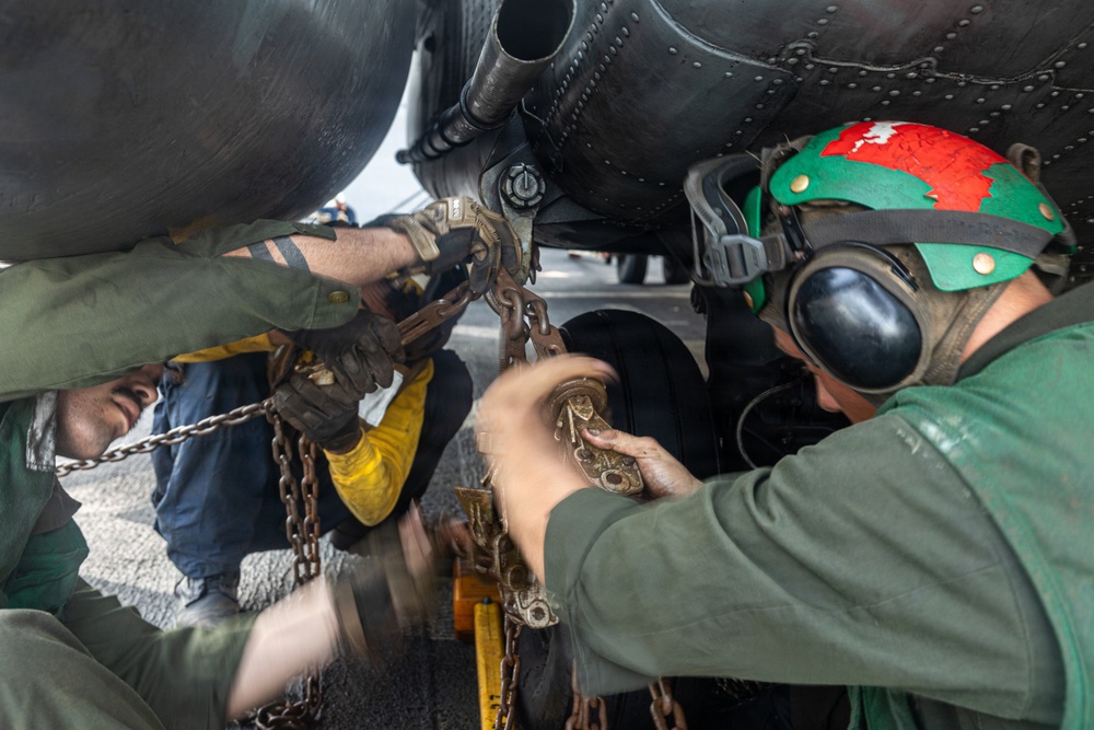 Military aviation deck crew members are working beneath an aircraft, handling tie-down chains and securing hardware on what appears to be a naval aircraft carrier flight deck. At least three personnel are visible, wearing color-coded gear indicative of their roles. The close-up angle captures the hands-on, physically demanding nature of aircraft chaining and securing operations.