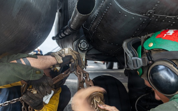 Military aviation deck crew members are working beneath an aircraft, handling tie-down chains and securing hardware on what appears to be a naval aircraft carrier flight deck. At least three personnel are visible, wearing color-coded gear indicative of their roles. The close-up angle captures the hands-on, physically demanding nature of aircraft chaining and securing operations.