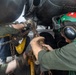 Military aviation deck crew members are working beneath an aircraft, handling tie-down chains and securing hardware on what appears to be a naval aircraft carrier flight deck. At least three personnel are visible, wearing color-coded gear indicative of their roles. The close-up angle captures the hands-on, physically demanding nature of aircraft chaining and securing operations.