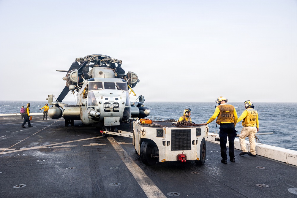 Naval personnel are repositioning a military helicopter on the flight deck of a warship at sea. A small tow tractor is being used to move the helicopter, with crew members in yellow jerseys and safety gear guiding the operation. The helicopter, marked with the number '22', has its rotor blades folded for deck storage.