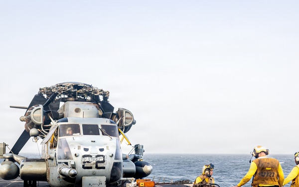Naval personnel are repositioning a military helicopter on the flight deck of a warship at sea. A small tow tractor is being used to move the helicopter, with crew members in yellow jerseys and safety gear guiding the operation. The helicopter, marked with the number '22', has its rotor blades folded for deck storage.