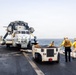 Naval personnel are repositioning a military helicopter on the flight deck of a warship at sea. A small tow tractor is being used to move the helicopter, with crew members in yellow jerseys and safety gear guiding the operation. The helicopter, marked with the number '22', has its rotor blades folded for deck storage.