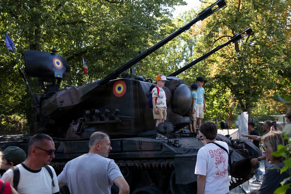 A military vehicle, likely a self-propelled anti-aircraft gun or artillery piece, is on public display in a park or outdoor exhibition setting. The vehicle bears Romanian roundel markings (red, yellow, blue concentric circles) and what appears to be a Romanian flag, suggesting this is a Romanian military open day or national holiday event. Civilians, including children climbing on the vehicle, are gathered around it in a relaxed, festive atmosphere.