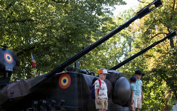A military vehicle, likely a self-propelled anti-aircraft gun or artillery piece, is on public display in a park or outdoor exhibition setting. The vehicle bears Romanian roundel markings (red, yellow, blue concentric circles) and what appears to be a Romanian flag, suggesting this is a Romanian military open day or national holiday event. Civilians, including children climbing on the vehicle, are gathered around it in a relaxed, festive atmosphere.