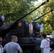 A military vehicle, likely a self-propelled anti-aircraft gun or artillery piece, is on public display in a park or outdoor exhibition setting. The vehicle bears Romanian roundel markings (red, yellow, blue concentric circles) and what appears to be a Romanian flag, suggesting this is a Romanian military open day or national holiday event. Civilians, including children climbing on the vehicle, are gathered around it in a relaxed, festive atmosphere.