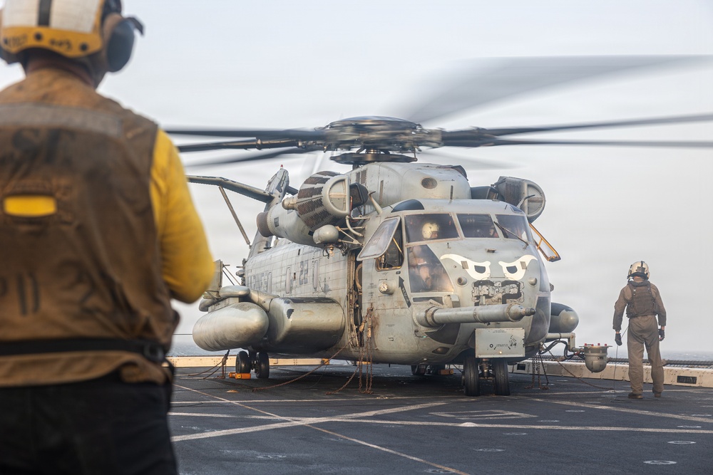 A large military helicopter, identifiable as a CH-53E Super Stallion, is positioned on the flight deck of a naval vessel with its rotors spinning. Two flight deck crew members in protective gear are visible — one in a yellow vest in the foreground and another in tan gear to the right. The scene depicts pre-flight or post-flight operations on what appears to be an amphibious assault ship.