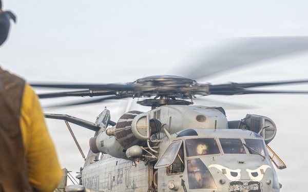 A large military helicopter, identifiable as a CH-53E Super Stallion, is positioned on the flight deck of a naval vessel with its rotors spinning. Two flight deck crew members in protective gear are visible — one in a yellow vest in the foreground and another in tan gear to the right. The scene depicts pre-flight or post-flight operations on what appears to be an amphibious assault ship.