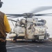 A large military helicopter, identifiable as a CH-53E Super Stallion, is positioned on the flight deck of a naval vessel with its rotors spinning. Two flight deck crew members in protective gear are visible — one in a yellow vest in the foreground and another in tan gear to the right. The scene depicts pre-flight or post-flight operations on what appears to be an amphibious assault ship.