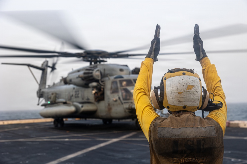 A naval flight deck crew member in a yellow jersey and helmet is directing a large military helicopter on what appears to be the deck of a naval vessel at sea. The crew member has both arms raised overhead in a standard aircraft handling signal. The helicopter, identifiable as a CH-53E Super Stallion, has its rotors spinning as indicated by motion blur.