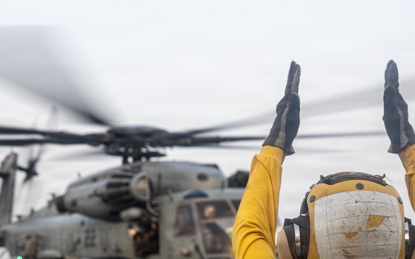 A naval flight deck crew member in a yellow jersey and helmet is directing a large military helicopter on what appears to be the deck of a naval vessel at sea. The crew member has both arms raised overhead in a standard aircraft handling signal. The helicopter, identifiable as a CH-53E Super Stallion, has its rotors spinning as indicated by motion blur.
