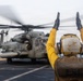 A naval flight deck crew member in a yellow jersey and helmet is directing a large military helicopter on what appears to be the deck of a naval vessel at sea. The crew member has both arms raised overhead in a standard aircraft handling signal. The helicopter, identifiable as a CH-53E Super Stallion, has its rotors spinning as indicated by motion blur.