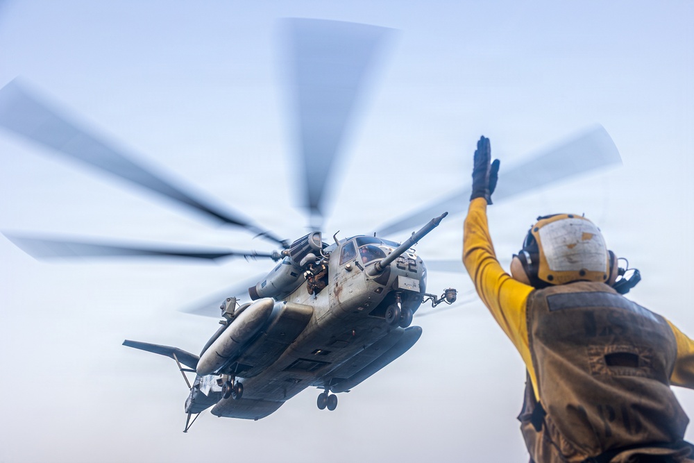 A military helicopter, appearing to be a CH-53E Super Stallion, is captured in a low-angle shot during a deck landing or departure operation. A flight deck crew member in yellow jersey and white helmet is giving hand signals to the aircraft. The rotor blades show significant motion blur, indicating active rotation at high speed.