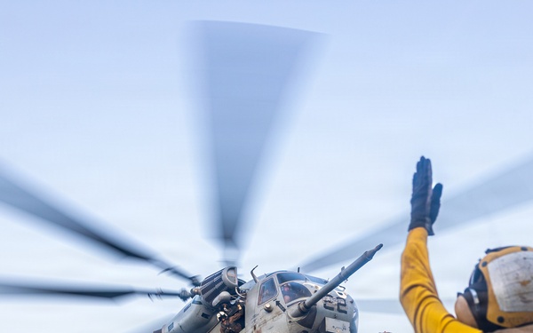 A military helicopter, appearing to be a CH-53E Super Stallion, is captured in a low-angle shot during a deck landing or departure operation. A flight deck crew member in yellow jersey and white helmet is giving hand signals to the aircraft. The rotor blades show significant motion blur, indicating active rotation at high speed.